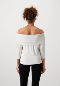 Off-the-shoulder topp i lett hvit stoff, tettsittende i midjen med 3/4-lange ermer. Glatt tekstur og subtile samlinger langs kantene.