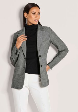 Grau melierter Herringbone-Blazer mit zwei Knöpfen, Reverskragen und langen Ärmeln, kombiniert mit einem schwarzen Rollkragenpullover und weißen Hosen.