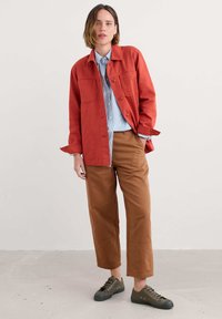 Roestkleurige jas over een lichtblauw gestreept shirt, gecombineerd met bruine cropped broek en olijfgroene sneakers met veters.