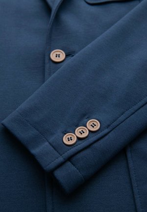Manica di un blazer blu navy con tre bottoni beige, che mostra dettagli di cucitura e la trama del tessuto.