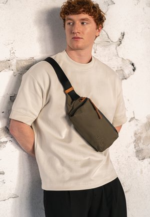 Borsa a tracolla verde oliva con cerniera, dettagli in pelle beige e tracolla nera regolabile, indossata across il torso da una persona in una camicia color crema.