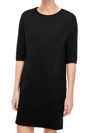 Kvinde iført en ensfarvet sort, kortærmet kjole, der går til knæene, med rund halsudskæring og sidelommer, stående mod en hvid baggrund.