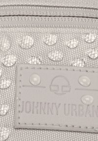 Detailní záběr na kapky vody na texturované tkanině s přišitou etiketou, na které jsou slabě vytištěny text a logo "Johnny Urban".