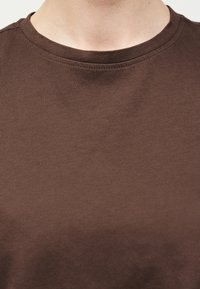 Κοντινό πλάνο ενός ατόμου που φοράει απλό καφέ μπλουζάκι με στρογγυλή λαιμόκοψη, εμφανίζοντας το πάνω μέρος του στήθους και τη λαιμόκοψη.