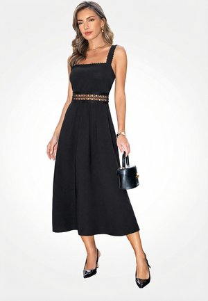 Vrouw in een mouwloze zwarte midi-jurk met kanten taille-detail, die een kleine zwarte handtas vasthoudt en zwarte hakken en gouden sieraden draagt.