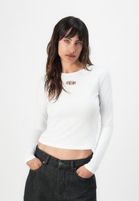 Crop top blanc à manches longues avec un col rond et un accent en métal argenté au niveau de la poitrine, porté avec un pantalon en denim foncé taille haute.