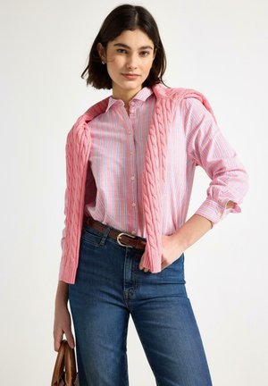 Donna che indossa una camicia a righe rosa e blu con un maglione rosa drappeggiato sulle spalle, jeans blu e tiene in mano una borsa marrone.