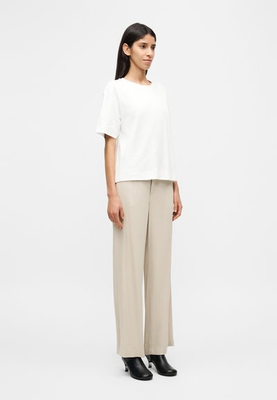 Λευκό κοντομάνικο πουκάμισο σε συνδυασμό με ανοιχτό μπεζ παντελόνι φαρδιάς γραμμής και μαύρα μποτάκια με τακούνι· το ύφασμα φαίνεται λείο με ελάχιστες σχεδιαστικές λεπτομέρειες.