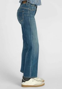 Jean en denim bleu à coupe droite, avec des poches arrière ornées d'un motif de couture distinctif, assortis à des baskets blanches.