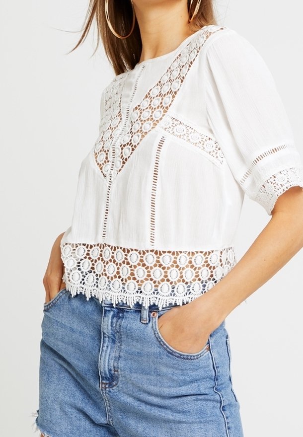 Crop top bianco con dettagli in pizzo intricati, dotato di scollatura rotonda e maniche corte. Abbinato a pantaloncini in denim a vita alta.