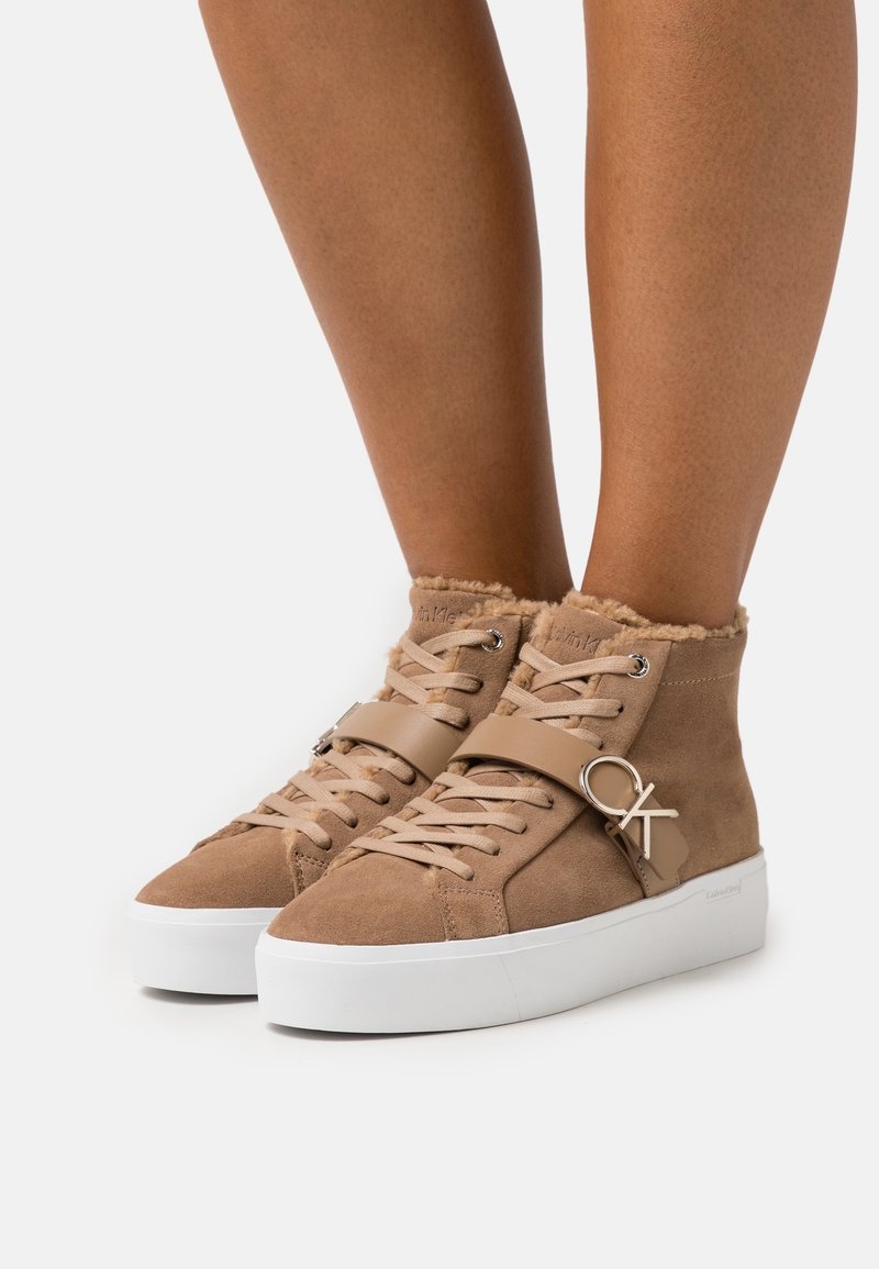 Sneakers alta in camoscio beige con colletto peloso, cinturino in cuoio beige, elementi in metallo argentato e suola in gomma bianca. Presentano un design di allacciatura classico.