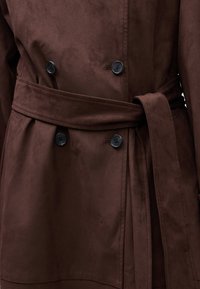 Trench coat in suede marrone con doppio petto, bottoni neri e cintura coordinata, che mette in mostra una trama liscia e un design sartoriale.