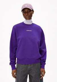 Paarse sweatshirt met ronde hals en geribbelde zoom, voorzien van witte logo-tekst. Gekomplimentiseerd met grijze jeans en een lichtpaarse pet.