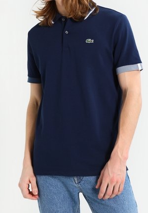 Marineblauw poloshirt gemaakt van katoen, met een klassieke kraag, korte opgerolde mouwen met grijze accenten, en een groen Lacoste-logo op de borst.