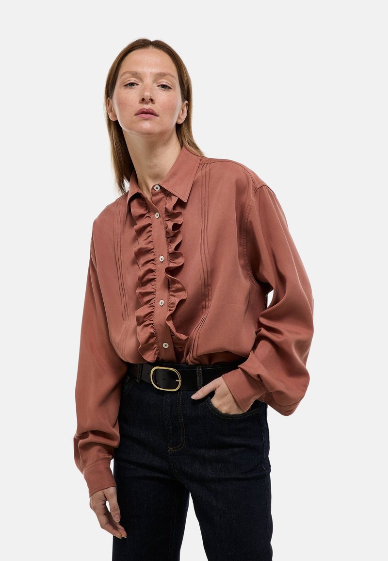 Blouse couleur rouille avec un détail froncé à l'avant, fermeture à boutons et col. Manches longues et tissu doux. Portée avec un jean sombre et une ceinture noire.