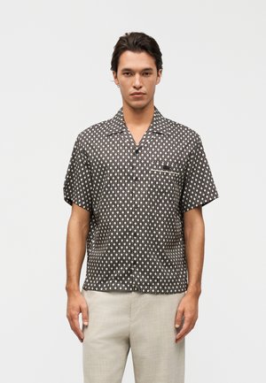 Homme portant une chemise noire à manches courtes avec un motif géométrique blanc et un pantalon beige clair, debout devant un fond uni.