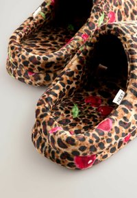 Leopard print slippers made of soft fabric, featuring red cherry patterns with green leaves on a tan background. Low, round, cushioned design.