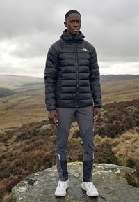 Jauns vīrietis melnā pūkainā jaku un pelēnās pārgājienu biksēs stāv uz akmens drēgnā, kalnainā ainavā.