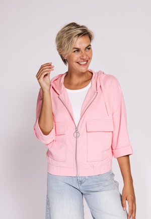 Smiling woman with short blonde hair wearing a pink zip-up jacket over a white top and light blue jeans, standing against a plain background.
