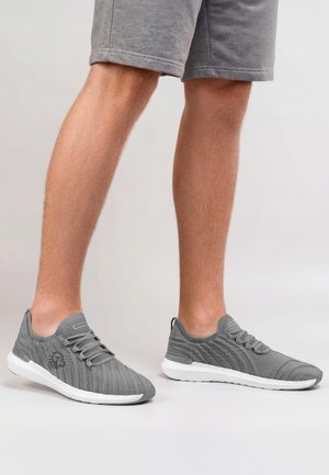 Mann trägt graue Stricksneaker mit weißen Sohlen und graue Shorts, steht vor einem hellgrauen Hintergrund.