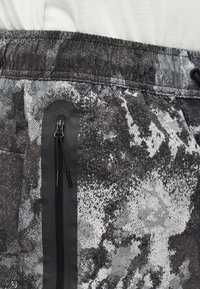 Κοντινό πλάνο σορτς από ύφασμα με σκούρο γκρι και λευκό σχέδιο, με μαύρη κάθετη τσέπη με φερμουάρ και ελαστική μέση.