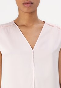 Blusa rosa pallido con scollatura a V, maniche corte e delicati bordi in pizzo sulle spalle; texture liscia e leggera lucentezza.