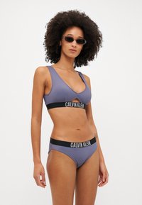Fialový žebrovaný bikinový kostým s výřezovým designem, vybavený černým elastickým páskem s logem "CALVIN KLEIN". Hladká textura, minimální design.