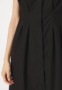 Μαύρο φόρεμα με διαρθρωτές πτυχές, λεία υφή και στενή σιλουέτα. Περιλαμβάνει μία διακριτική ραφή στη μέση μπροστά.