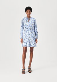 Blaues Blumenkleid mit langen Ärmeln, Kordelzug-Ausschnitt und gerafftem Saum. Es zeichnet sich durch einen leichten Stoff mit strukturiertem Muster aus. Schwarze Sandalen werden präsentiert.
