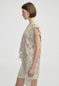 Haut en crochet beige avec des motifs floraux et des découpes sur les côtés, à coupe décontractée et sans manches, porté sur une robe légère.