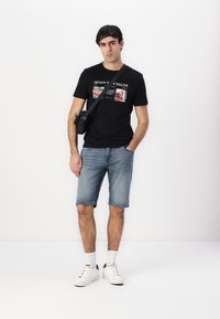 Svart grafisk t-skjorte med forskjellige bilder og tekst, kombinert med lys blå denimshorts og hvite sneakers med svarte detaljer.