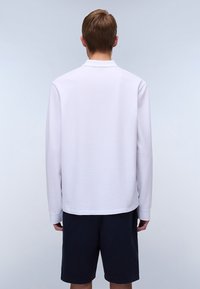 Homme aux cheveux bruns courts portant une chemise blanche à manches longues et un short noir, de dos, sur un fond gris clair.