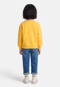 Παιδί με κοντά σκούρα μαλλιά, φοράει κίτρινο φούτερ, γυρισμένο τζιν παντελόνι, ριγέ κάλτσες και λευκά αθλητικά παπούτσια, στέκεται γυρισμένο με την πλάτη σε λευκό φόντο.