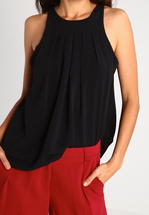 Vrouw die een mouwloze zwarte geplooide blouse draagt, ingestopt in een hoge taille rode broek met handen in de zakken.