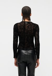 Άτομο με κοτσίδες cornrow που φοράει διάφανο μαύρο τοπ με μοτίβο φίδι και μαύρο δερμάτινο παντελόνι, στέκεται με την πλάτη στην κάμερα.