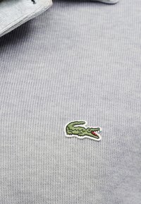 Tissu tricoté gris clair avec un petit logo crocodile vert brodé près du bord du col.
