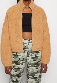 Veste en polaire beige, confortable, avec un col haut et une fermeture éclair. Présente des manches amples et une surface texturée. Associée à un pantalon camouflage.