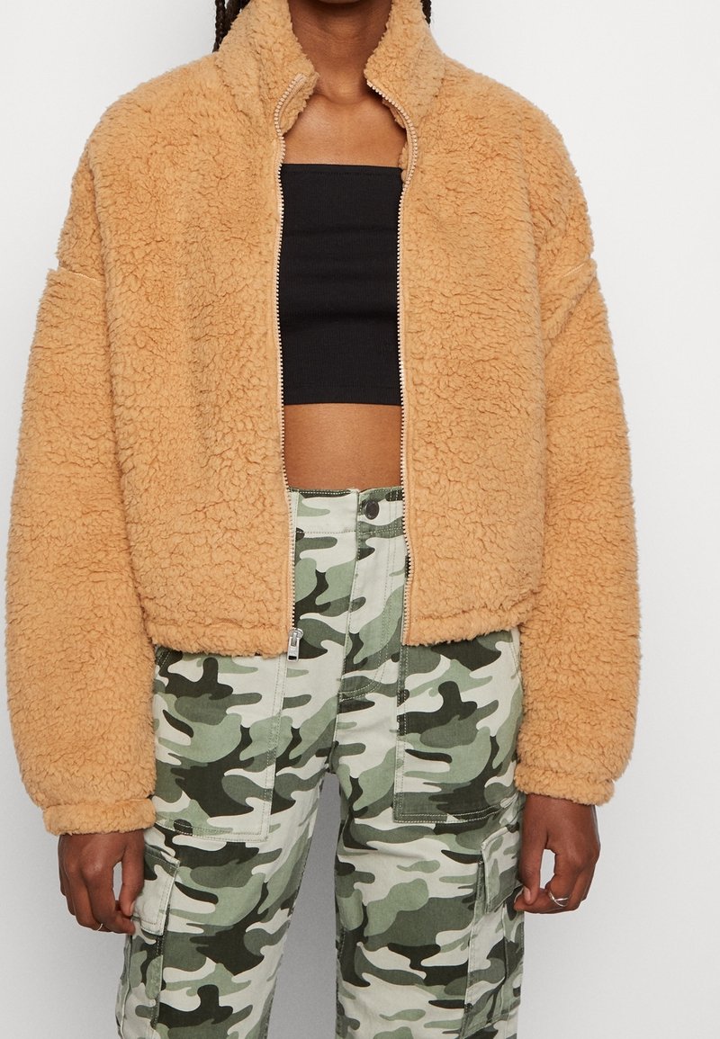 Veste en polaire beige, confortable, avec un col haut et une fermeture éclair. Présente des manches amples et une surface texturée. Associée à un pantalon camouflage.