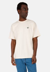 Cremefarbenes Baumwoll-T-Shirt mit Rundhalsausschnitt und kurzen Ärmeln. Verfügt über ein kleines grünes Logo auf der linken Brust und hat eine lockere Passform.