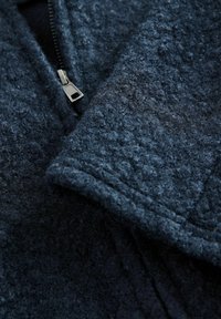 Tmavomodrý, textúrovaný fleecový materiál so zipsom; okraj goliera má hladký povrch. Viditeľné prešívanie po celom povrchu.