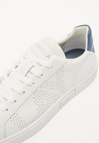 White leather sneaker with perforated pattern, rounded toe, and white laces. Features a grey-blue heel accent and textured rubber sole.