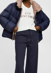 Chaqueta de plumas azul marino con cuello beige, camiseta blanca con la impresión "UNIRSE" y pantalones azul marino con cintura elástica.