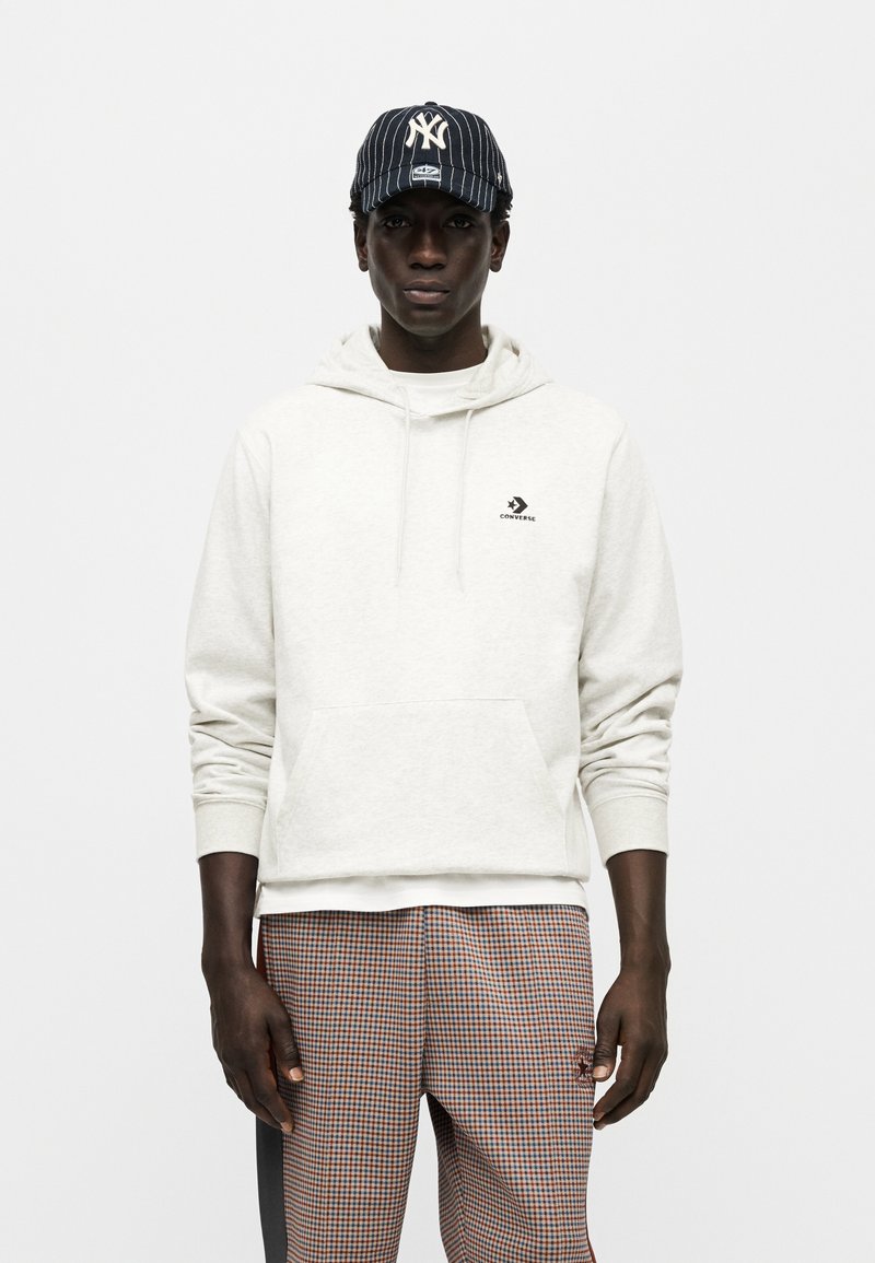 Sweat à capuche en polaire gris clair avec poche kangourou et petit logo Converse noir, associé à un pantalon à carreaux et une casquette marine à fines rayures.