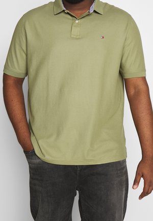 Mann trägt ein olivgrünes Polohemd mit kurzen Ärmeln und kleinem Logo sowie ausgewaschene schwarze Jeans, eine Hand in der Tasche, die andere entspannt an der Seite.