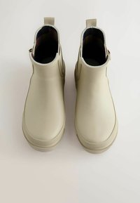 Bottes de cheville en caoutchouc beiges avec une texture lisse, bout rond et tirant. L'intérieur présente une doublure à carreaux. Design élégant et moderne.