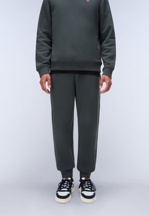 Mann trägt dunkelgrüne Jogginghose und Sweatshirt mit schwarzen und weißen Sneakers und steht vor einem einfarbig hellen Hintergrund.