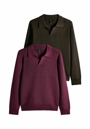 Junger Mann mit lockigem Haar, der einen burgunderfarbenen Pullover über einem dunkel karierten Hemd und hellblauen Jeans trägt, die Hände in den Taschen.