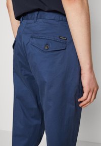 Marineblauwe katoenen broeken met een gladde textuur, bevatten een achterzak met een knoopsluiting en een klein merklabel aan de zijkant.