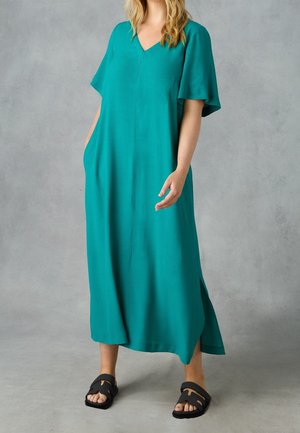 Vrouw die een losse teal midi-jurk draagt met V-hals en korte mouwen, staand met één hand in de zak en zwarte slip-on sandalen.