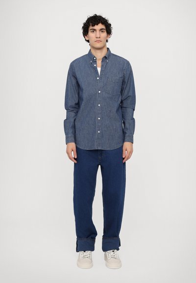 Camicia in denim blu scuro con bottoni, maniche lunghe e taschino sul petto, abbinata a jeans blu larghi e sneaker bianche.